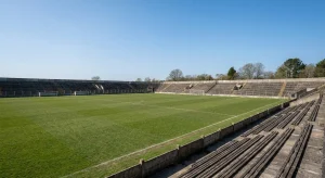 A small traditional sports stadium with a green football pitch and empty spectator stands under a clear sky