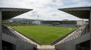 A wide shot of a soccer stadium with a green pitch, concrete grandstands, and two large overhanging roofs. The