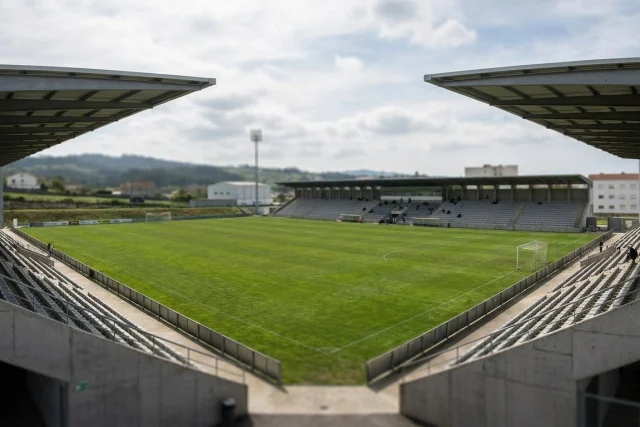 A wide shot of a soccer stadium with a green pitch, concrete grandstands, and two large overhanging roofs. The